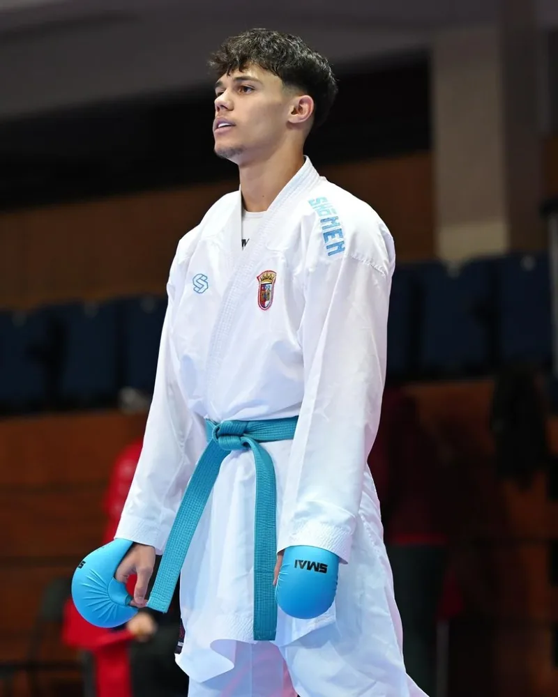 Santiago Gonçalves em pé no pavilhão, com kimono Shomen, cinto azul e luvas de kumite